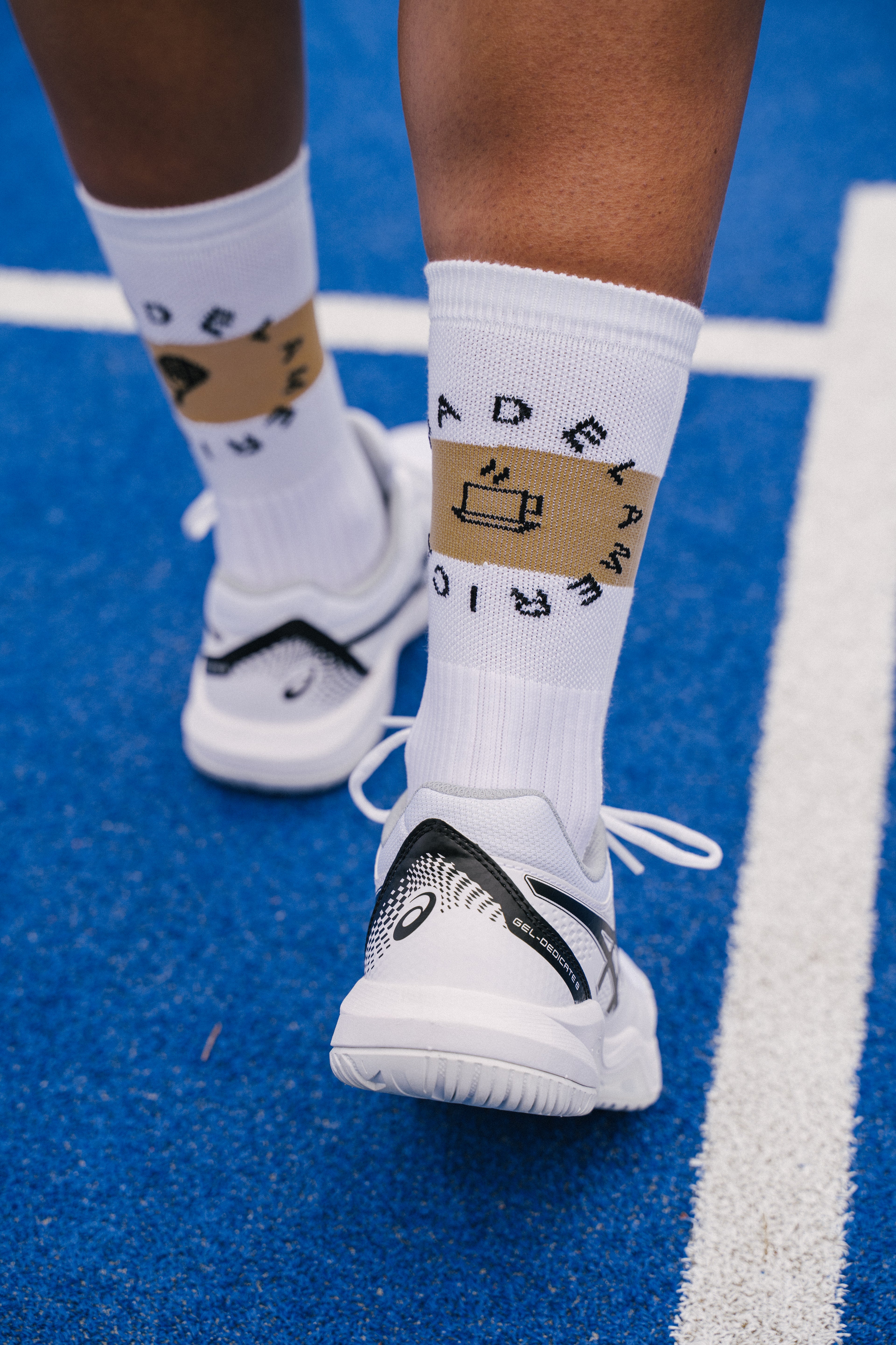 White padel socks with a beige horizontal stripe on the cuffs, and black text reading 'PADEL AMERICANO' arranged in a circle, worn with white and black padel shoues on a blue padel court.