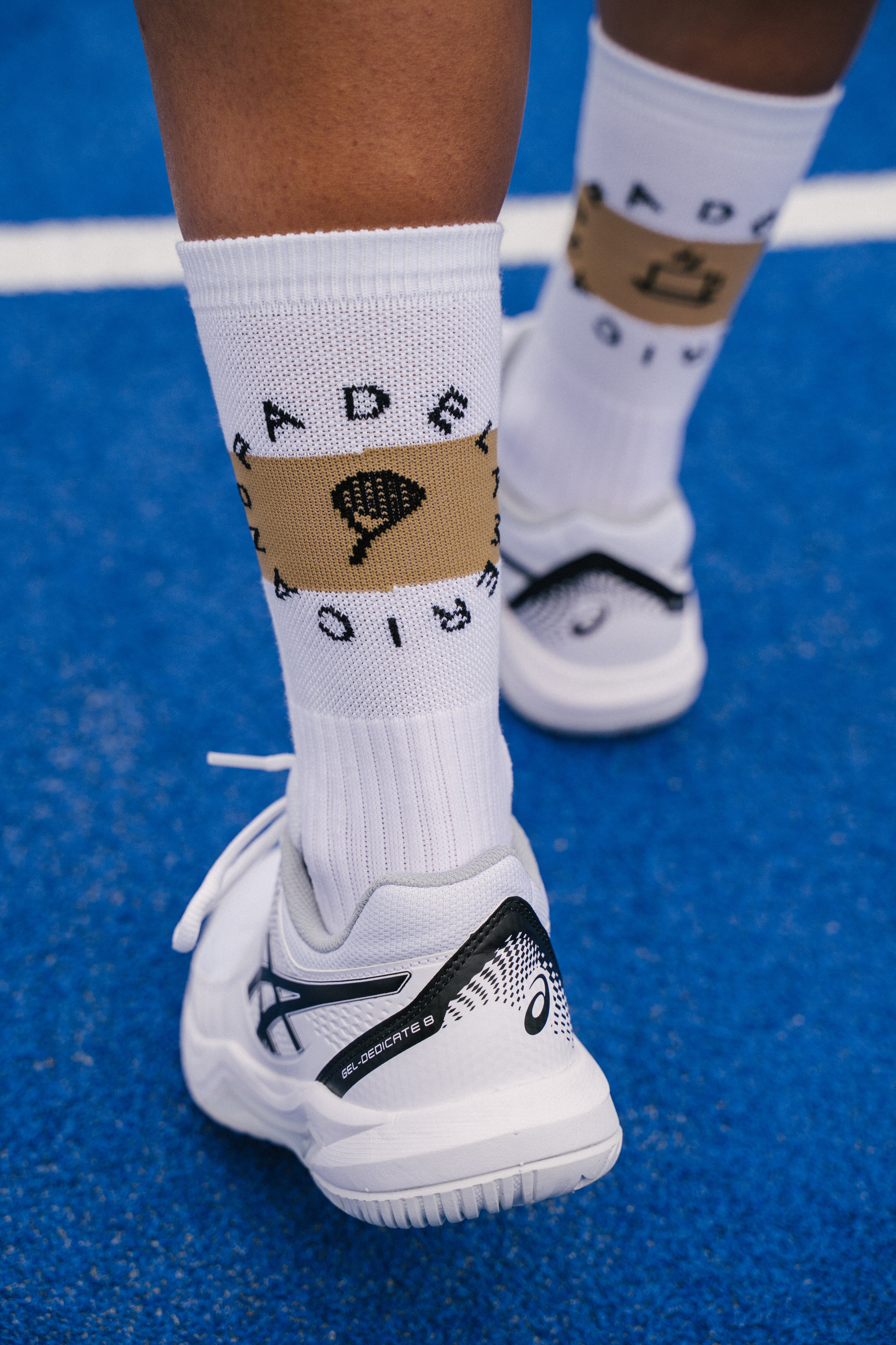 White padel socks with a beige band featuring a padel racket graphic and the text 'PADEL' and 'AMERCIANO', worn with white padel shoes on a blue padel court.