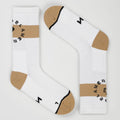 A pair of white padel socks with beige toe and heel sections, a beige horizontal stripe on the cuffs, and black text reading 'PADEL AMERICANO' arranged in a circle. One sock is upright and the other is angled diagonally, set against a clean, light gray background.