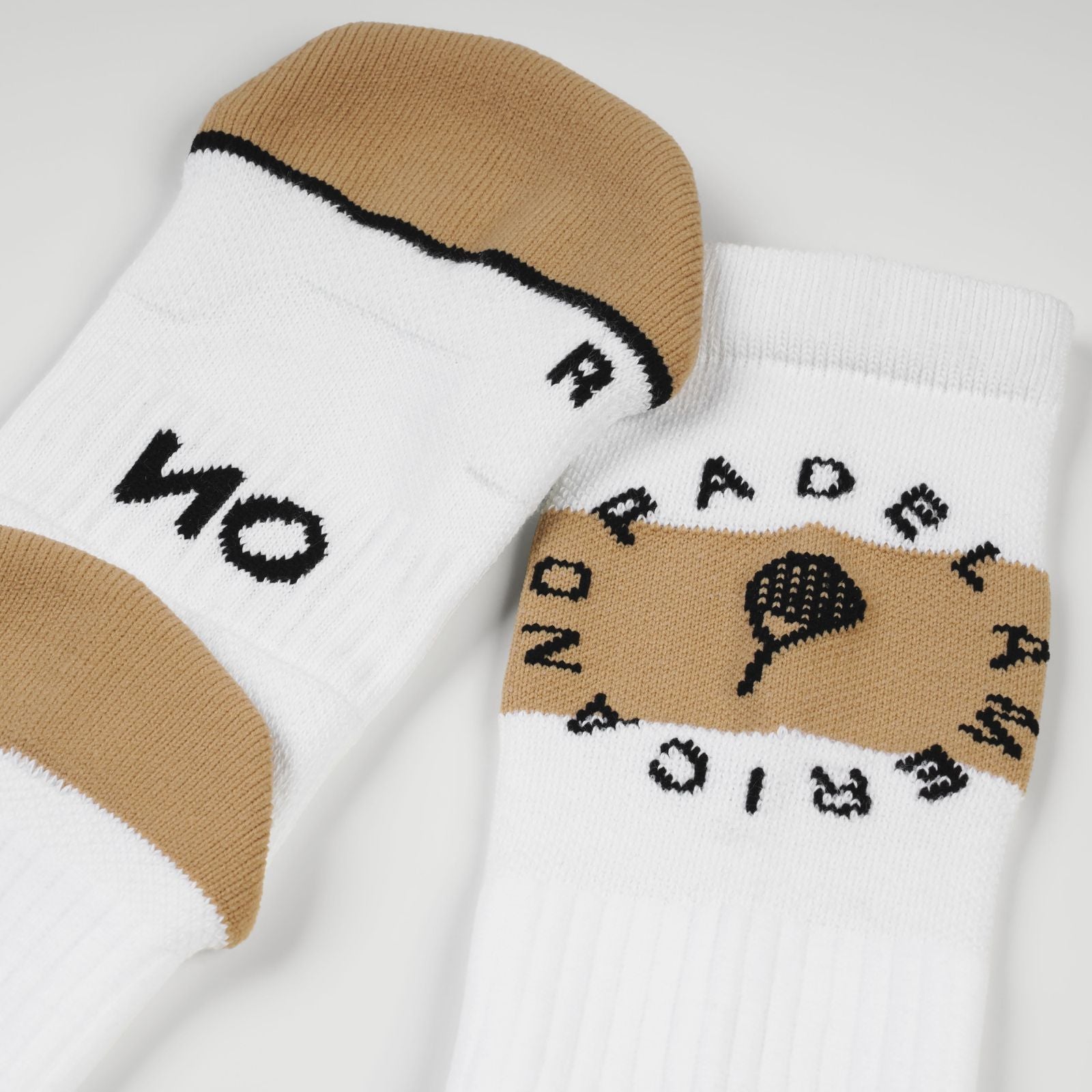 A pair of white padel socks with beige toe and heel sections, featuring black knit lettering and a padel racket graphic. One sock displays 'NO' and the other shows 'PADEL AMERICANO' arched around a padel racket motif, set against a clean white background.