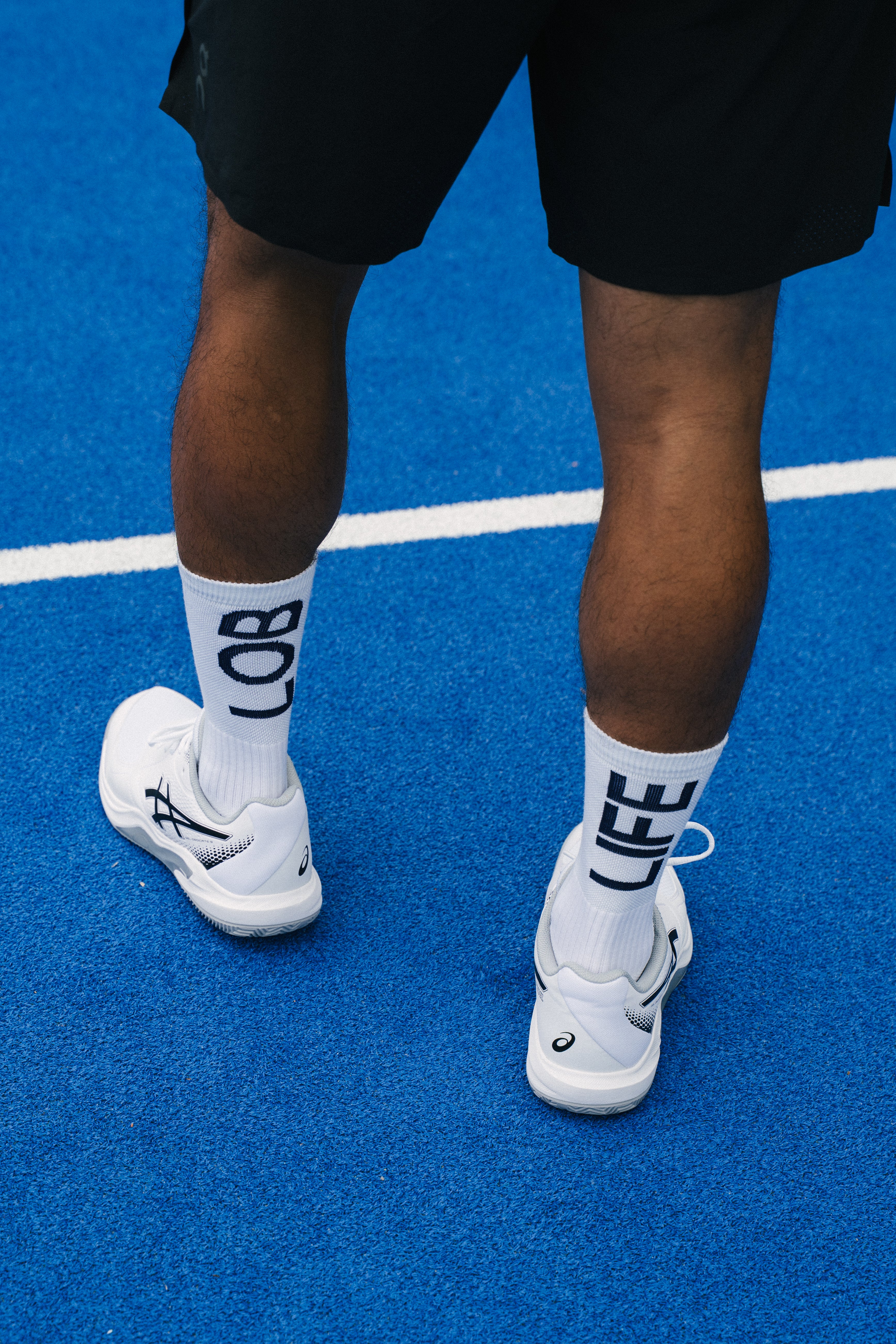 A close-up of a person's legs from the waist down, wearing black padel shorts, white padel socks with a navy blue statement 'LOB LIFE', and white padel shoes on a blue padel tennis court.