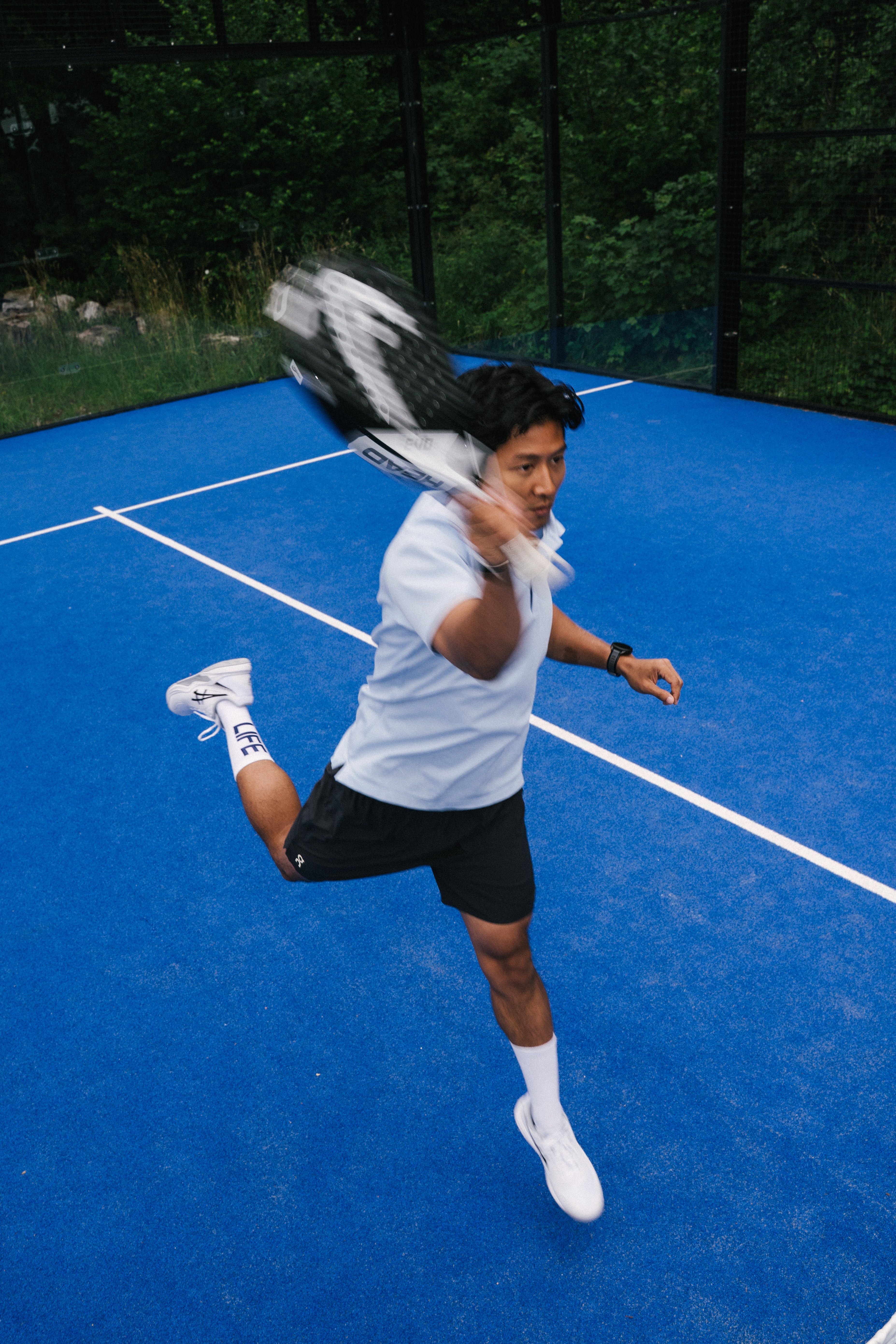 A padel player in athletic wear—a white t-shirt, black shorts, white padel socks, and white padel shoes—is mid-swing on a blue padel court, holding a padel racket. The background features a black fence and green trees.