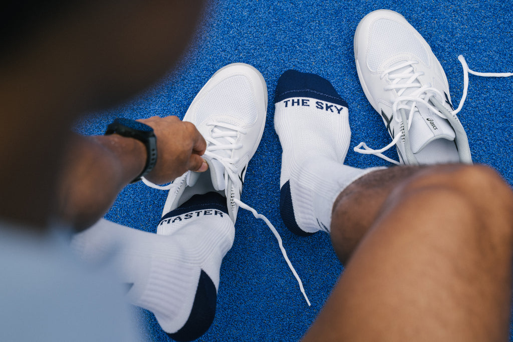 A close-up lifestyle shot of a person tying the laces of a white padel shoe while wearing white padel socks with navy blue heels and toes. One sock features the text 'MASTER' and the other shows 'THE SKY'.