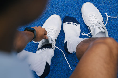 A close-up lifestyle shot of a person tying the laces of a white padel shoe while wearing white padel socks with navy blue heels and toes. One sock features the text 'MASTER' and the other shows 'THE SKY'.