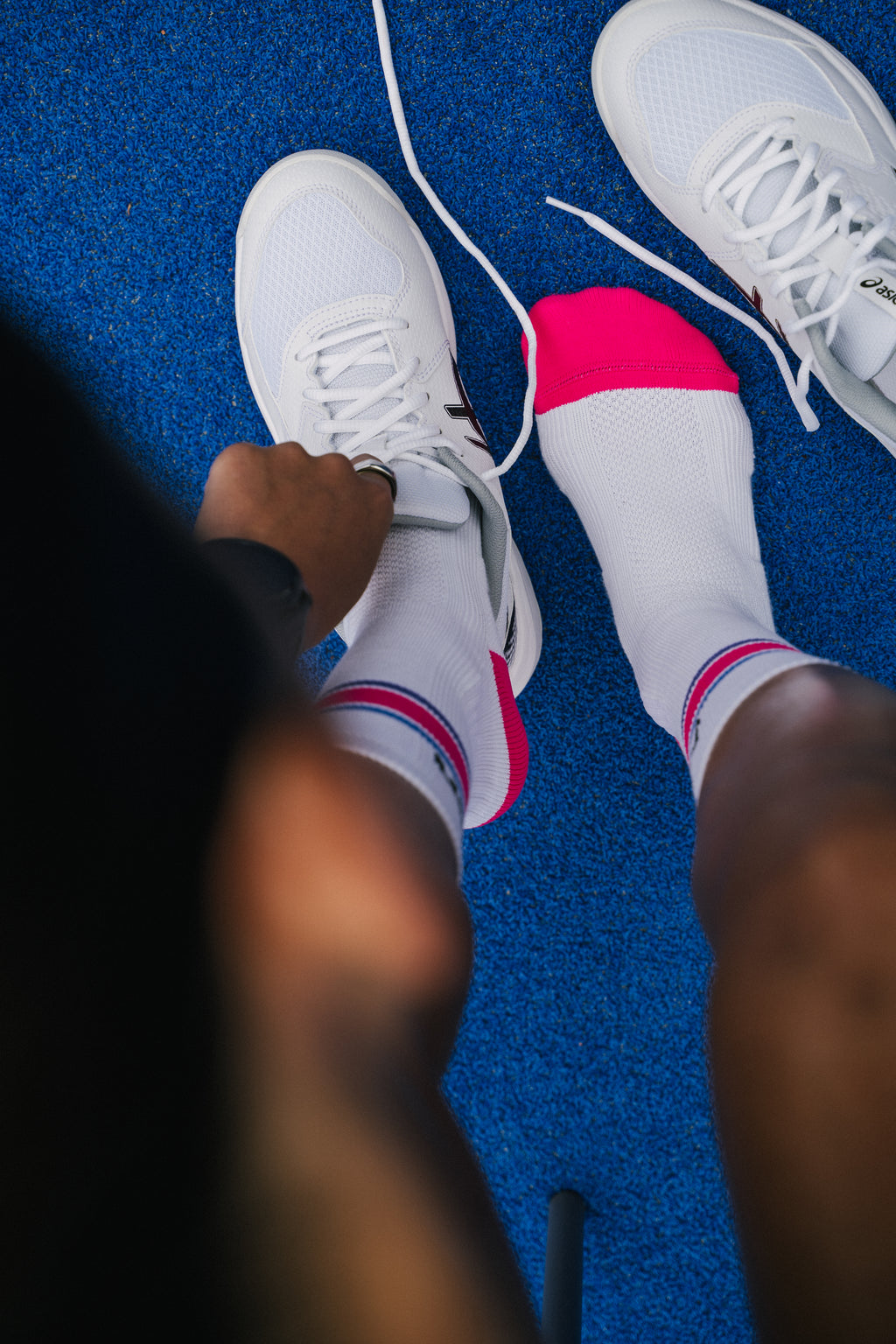 White padel socks with bright pink toes and blue pink stripes, worn with white padel shoes on a textured blue padel court floor, viewed from above.
