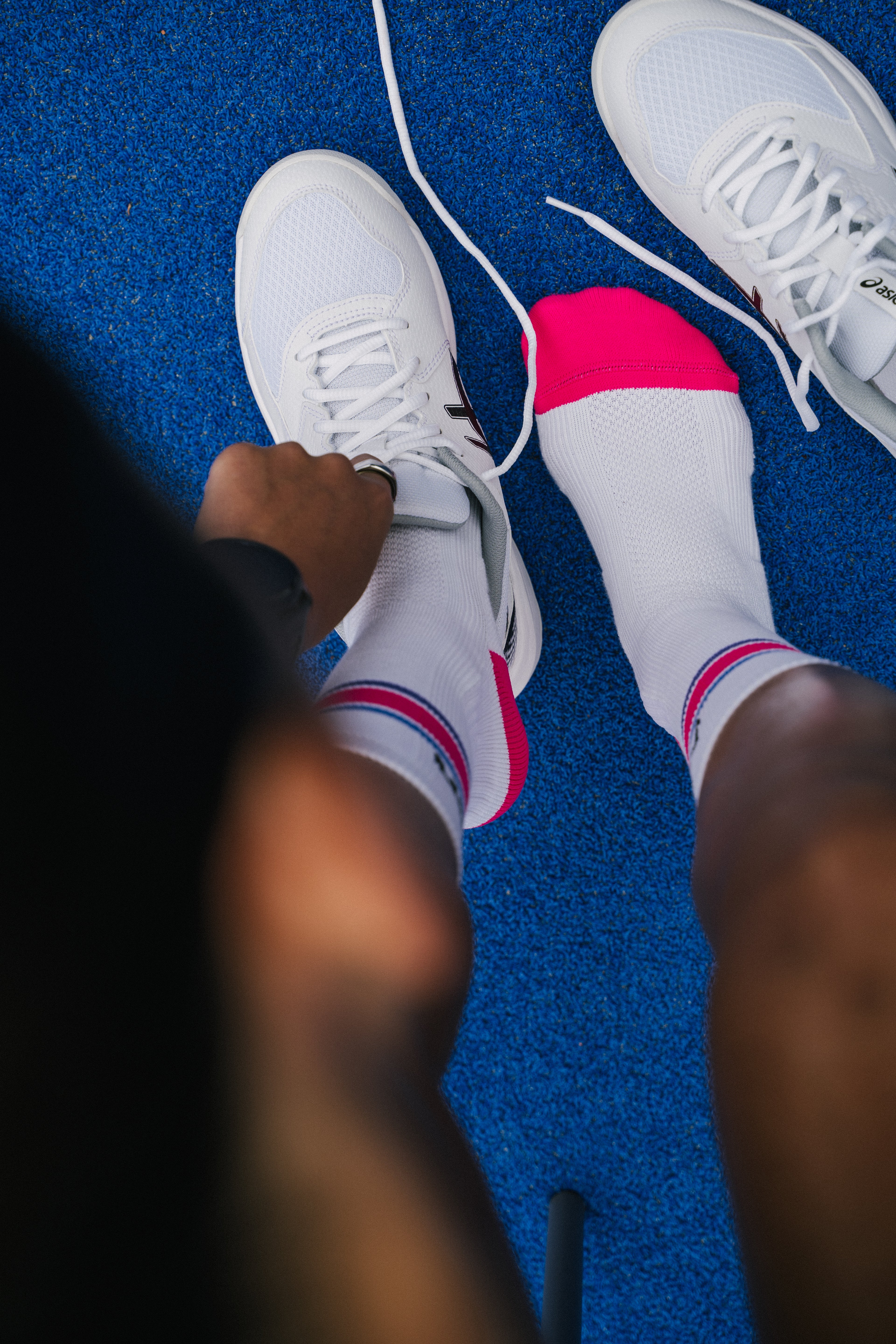 White padel socks with bright pink toes and blue pink stripes, worn with white padel shoes on a textured blue padel court floor, viewed from above.