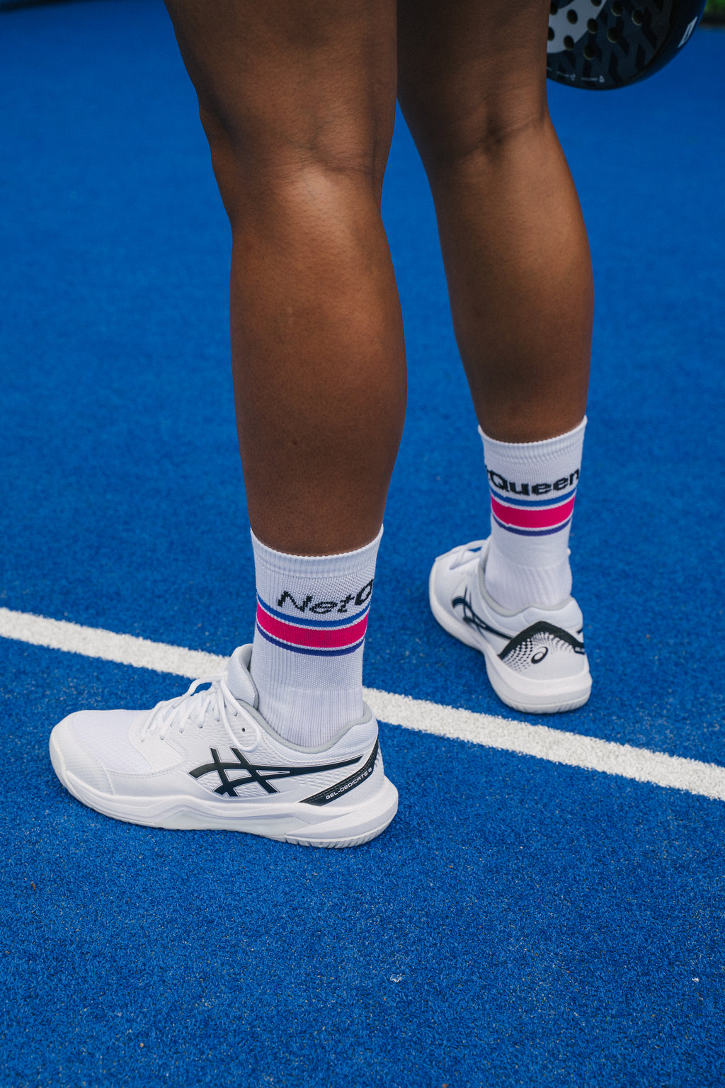 A close-up lifestyle shot of a padel player's legs from behind, wearing white padel shoes, white socks with pink and blue stripes and 'Net Queen' branding, standing on a blue padel court.