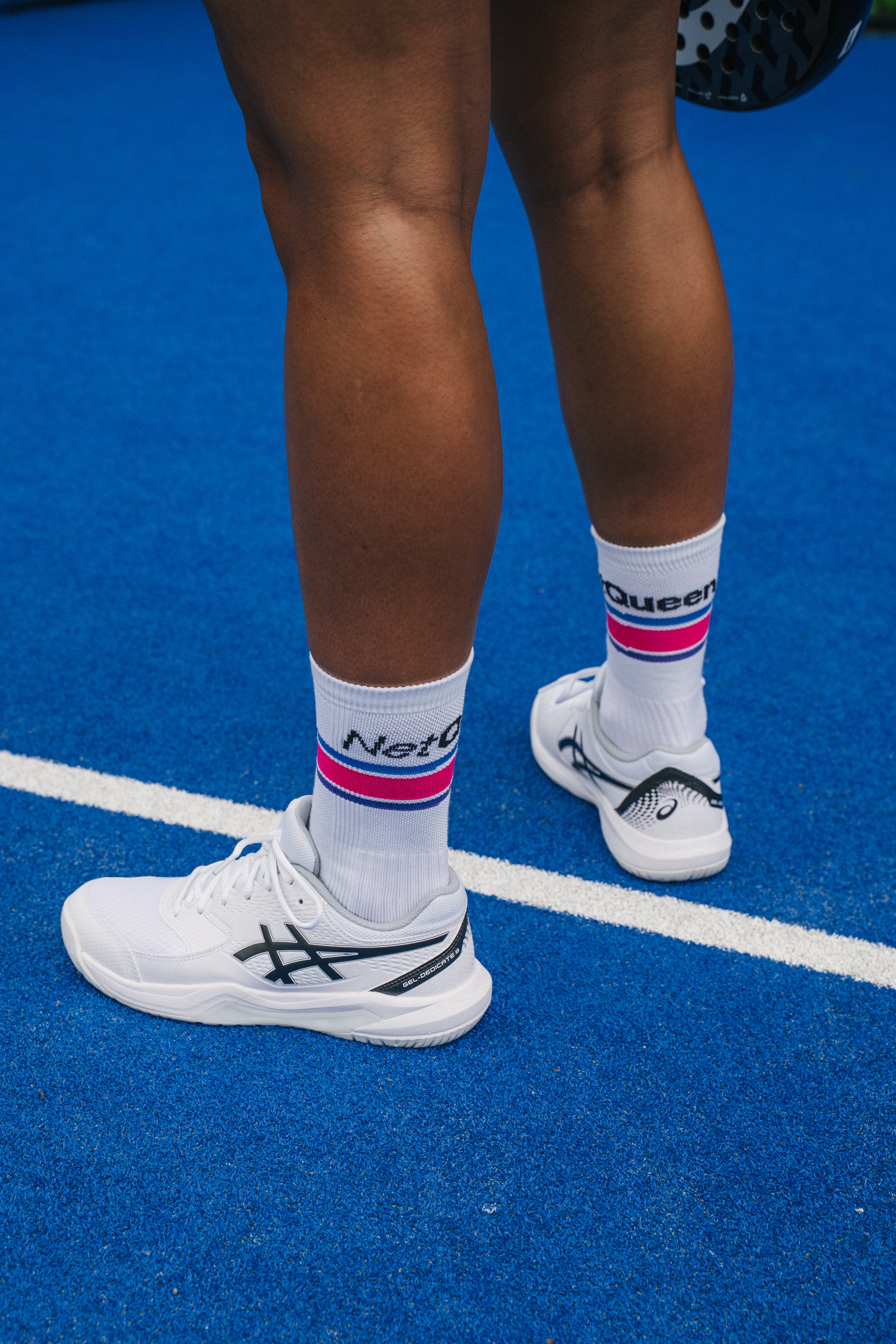 A close-up lifestyle shot of a padel player's legs from behind, wearing white padel shoes, white socks with pink and blue stripes and 'Net Queen' branding, standing on a blue padel court.