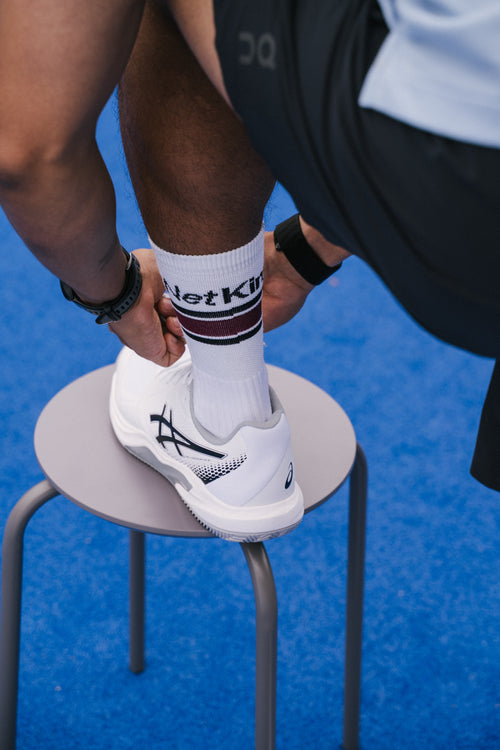 A close-up lifestyle shot of a person tying the ankle strap of a white padel shoe while wearing a white NoTennis padel sock with 'Net King' printed on it.