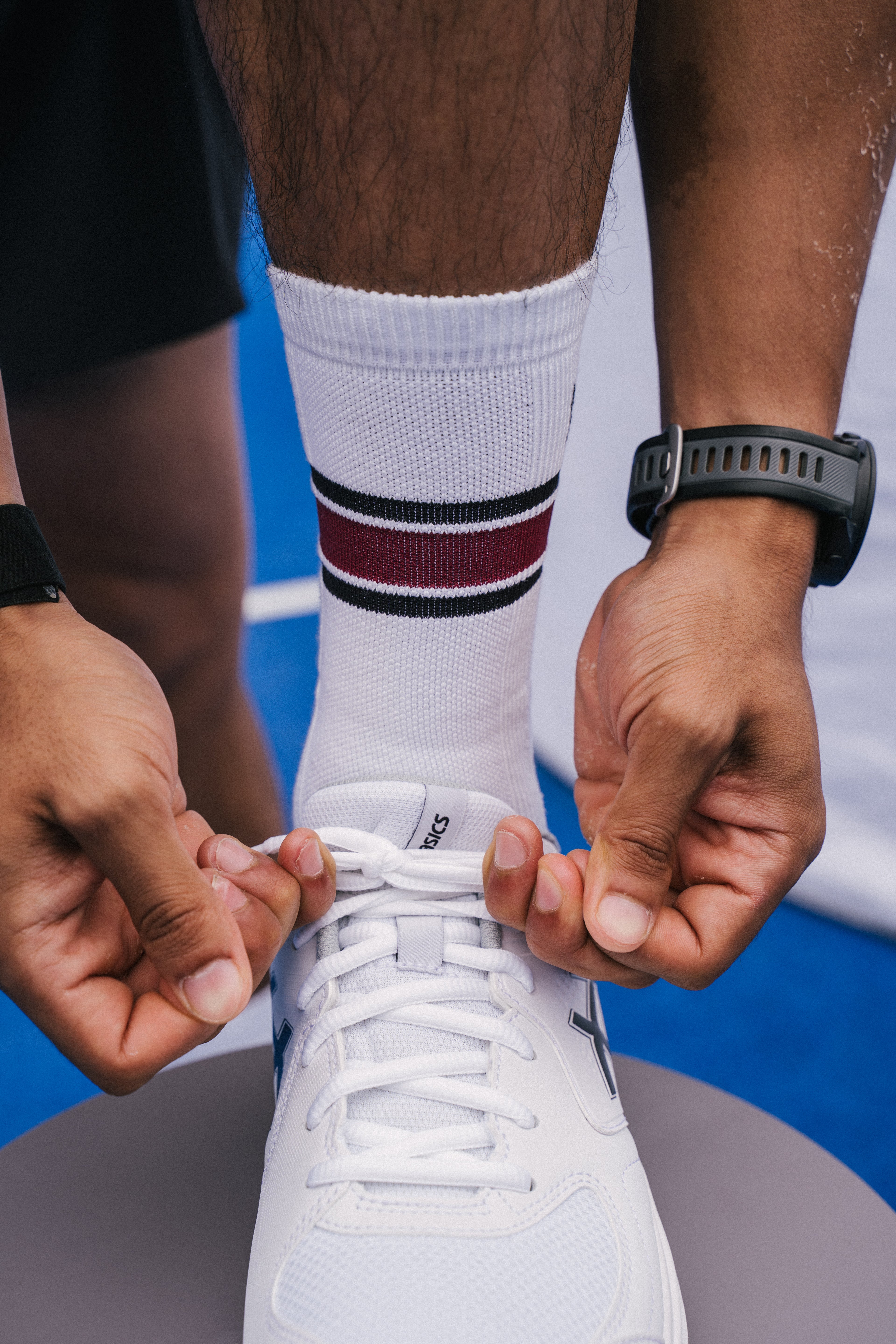 A close-up of a person tying the laces of a white padel shoe, wearing a white NoTennis padel court sock with black and bordeaux stripes, on a blue padel court surface.