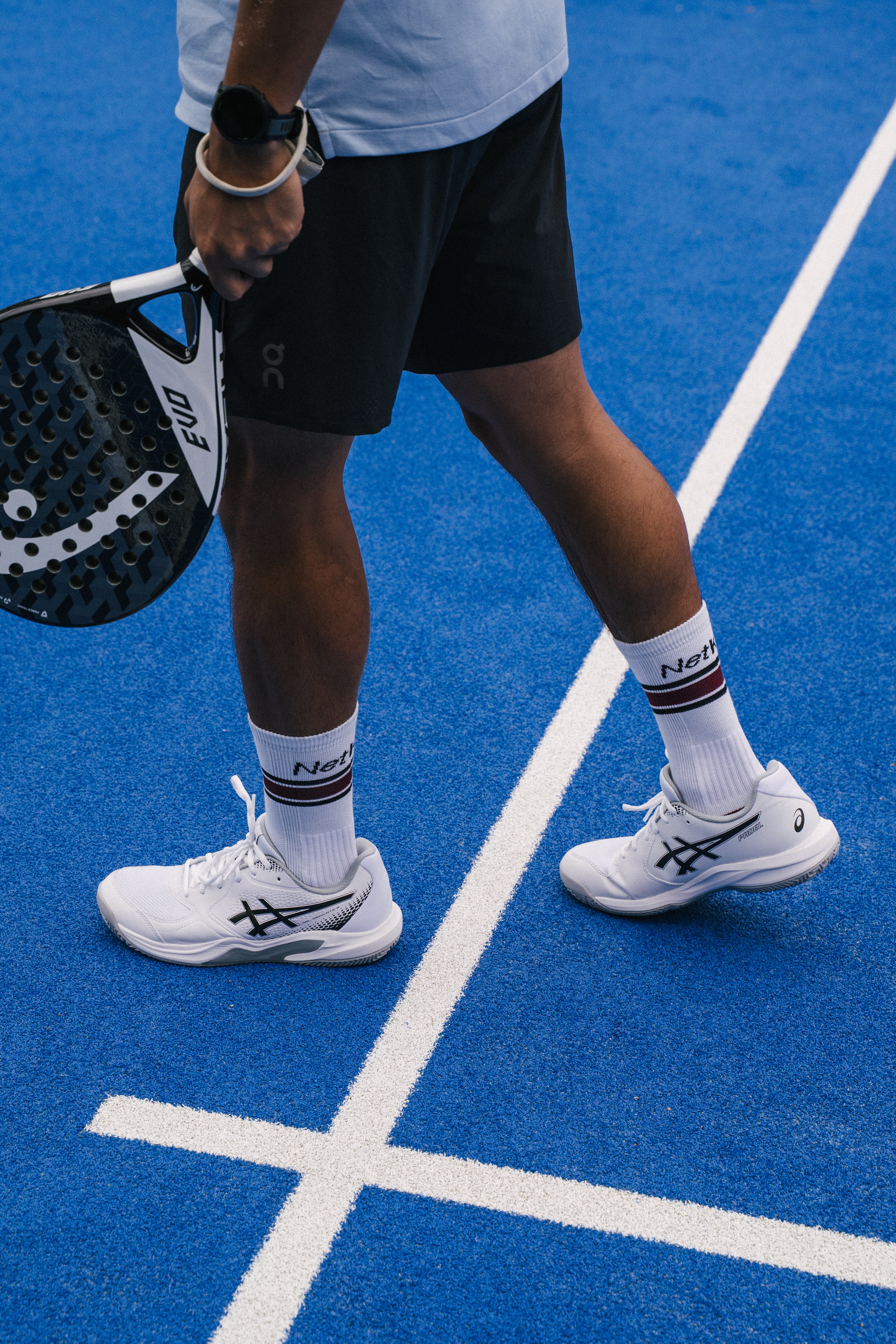 A close-up lifestyle shot of a padel tennis player on a blue padel court, wearing white padel shoes, white padel socks with red and black stripes, and black padel shorts, holding a padel racket.